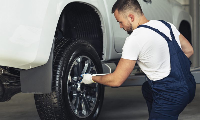 A man in overalls works on a tire of a white car, highlighting his expertise in vehicle repair and maintenance