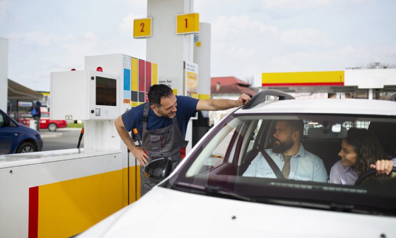 A man and woman refuel their car at a gas station, surrounded by fuel pumps and a clear blue sky.